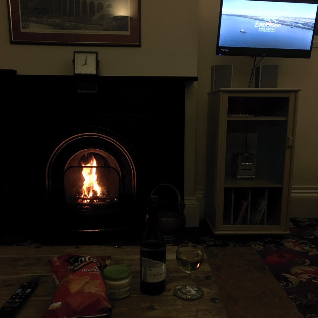 Cottage living room in the evening. A roaring fire in the fireplace. TV is showing the Eurovision song contest. On the table is a bottle of wine with a glass next to it, and a bag of Doritos with dip.