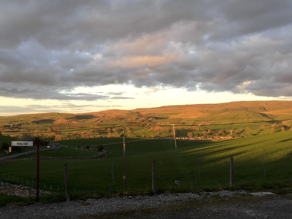 View of the countryside from the rear of Kirkby Stephen station. The hills are bathed in a golden glow from the setting sun. The station's "way out" sign is visible in the bottom-left corner.