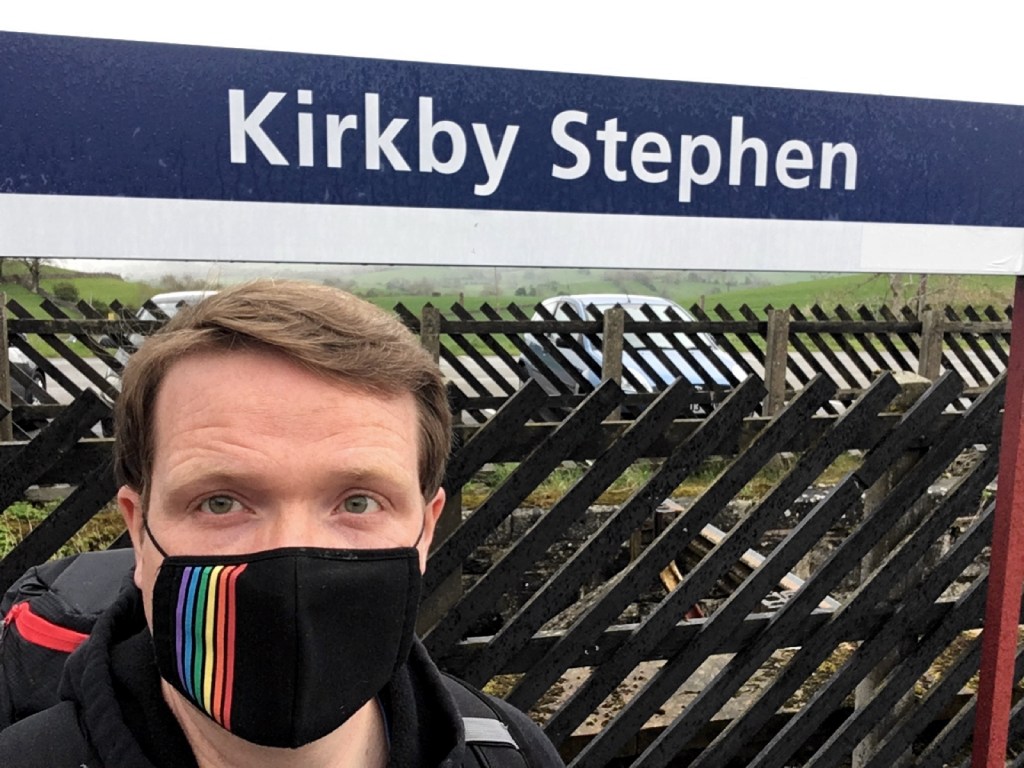 Robert standing in front of platform sign at Kirkby Stephen