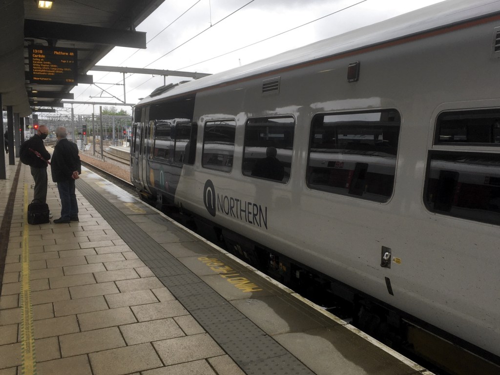 Northern class 158 train in a platform at Leeds. A couple of intending passengers stand on the platform waiting. Information display indicates this is the 1318 to Carlisle