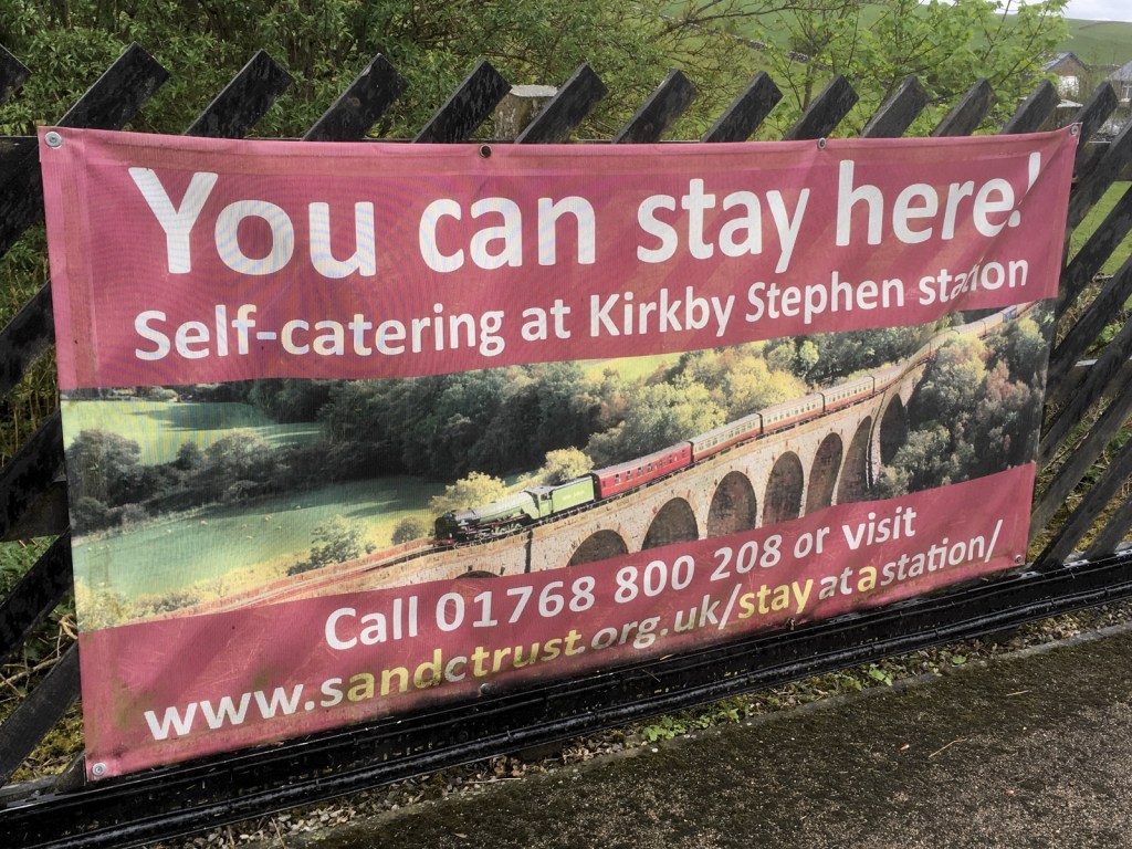 Banner attached to fence at Kirkby Stephen station. "You can stay here! Self-catering at Kirkby Stephen station" with a photo of a steam train crossing a viaduct.